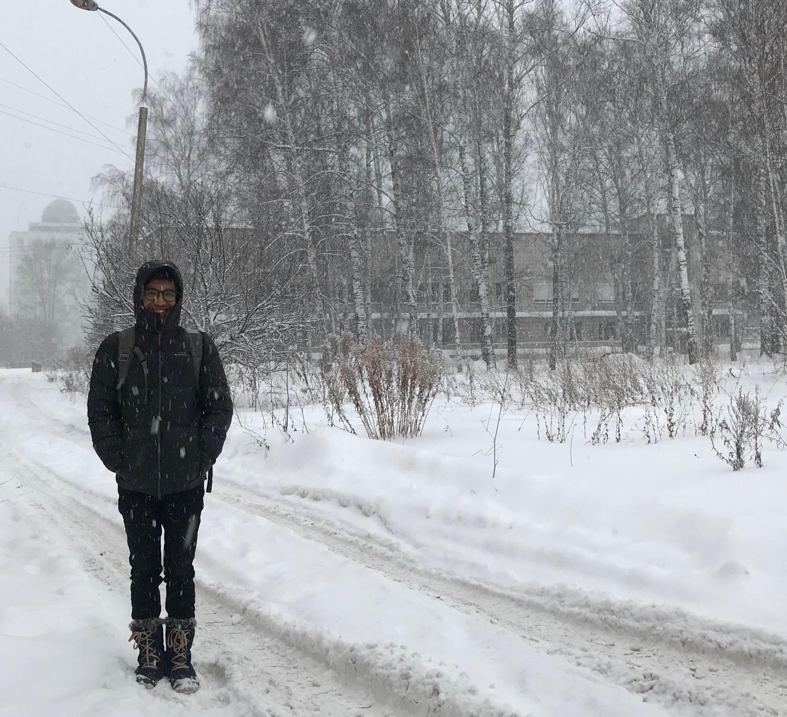 A picture of me. I'm dressed in a skiing jacket, warm boots, and a backpack. I'm standing on a snow-covered road while more snow falls around me. In the background you can trees and shrubs blanketed in snow. My face is mostly covered by the hood of my jacket, and all you can see is my eyes, nose, and mouth peeking out. Against all odds, I'm smiling.