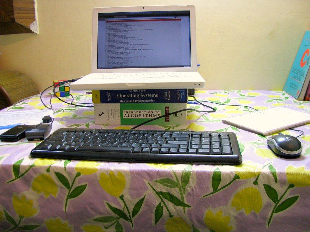 An improvised laptop stand built with books. A white plastic MacBook is placed on top of 'Operating Systems' and 'Introduction to Algorithms'. A keyboard and mouse connected to the laptop are placed on the table in front of the books. The tablecloth has a floral pattern with yellow flowers and green leaves.