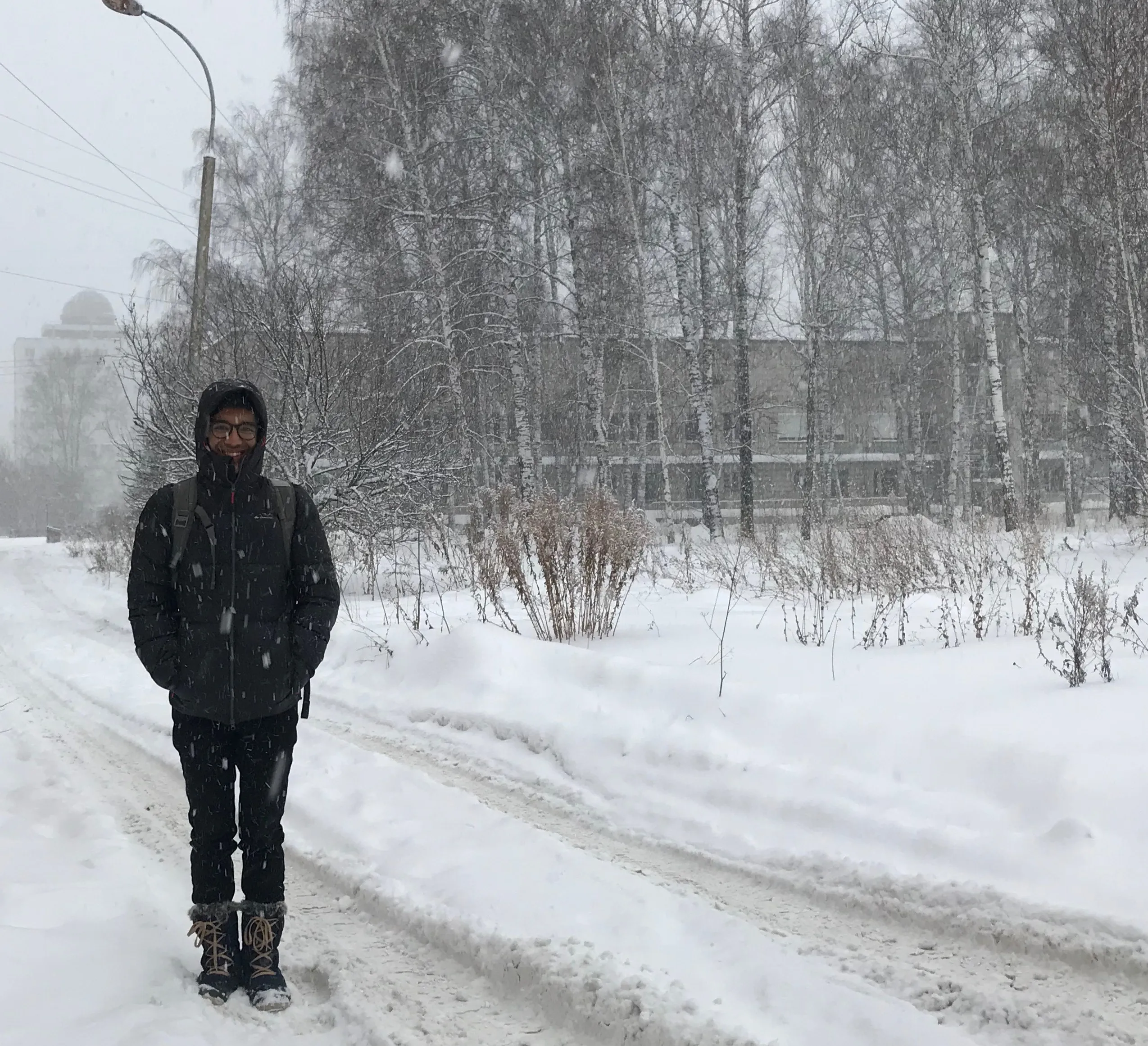 A picture of me. I&#x27;m dressed in a skiing jacket, warm boots, and a backpack. I&#x27;m standing on a snow-covered road while more snow falls around me. In the background you can trees and shrubs blanketed in snow. My face is mostly covered by the hood of my jacket, and all you can see is my eyes, nose, and mouth peeking out. Against all odds, I&#x27;m smiling.