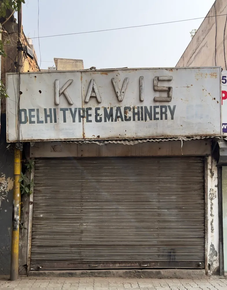 A shuttered type &#x26; machinery store near Jhandenwalan in Delhi. Old and weathered storefront sign reads Kavis Delhi Type &#x26; Machinery. The metal sign is rusted and worn, the paint peeling off it, and the rolling shutter is pulled down. The building shows signs of age and disrepair, with peeling paint and vegetation growing on the side.